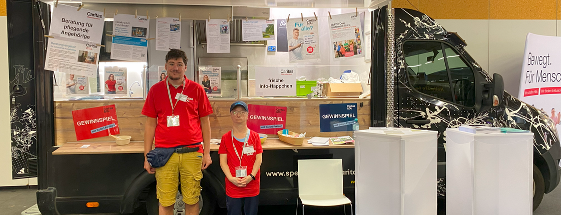 Ein Mann und eine Frau stehen vor einem Food Truck der mit Infomaterial dekoriert ist.