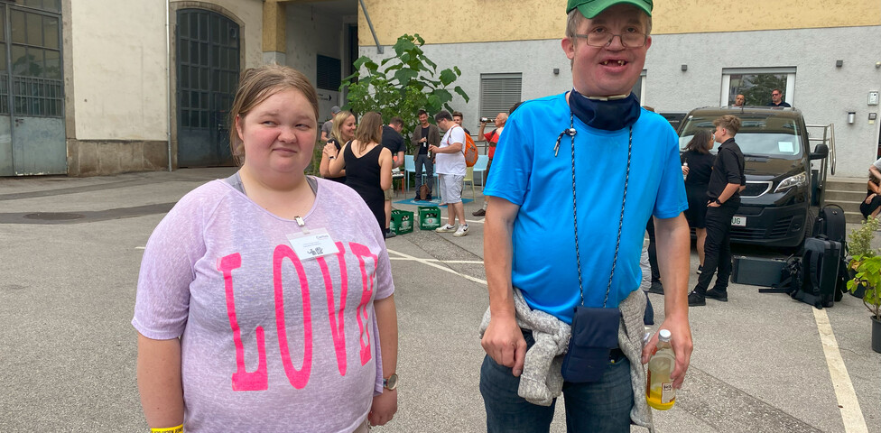 Eine Frau in einem rosa Shirt und ein Mann in einem blauem Shirt stehen vor einem Gebäude. 