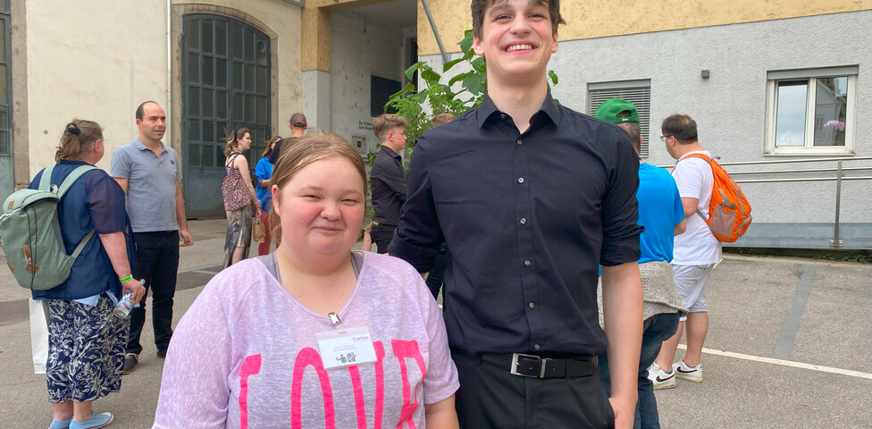 Eine Frau in rosa Shirt und ein Mann in schwarzem Hem stehen vor einem Gebäude. 